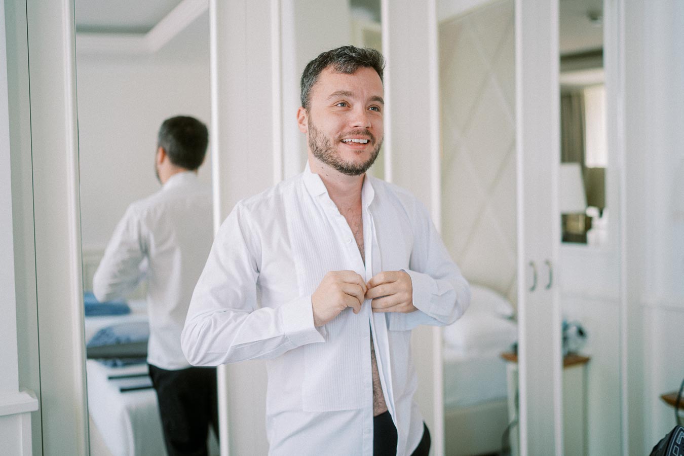 Man smiling while buttoning up a white shirt in a bright, modern bedroom with mirrors and a neatly made bed in the background.