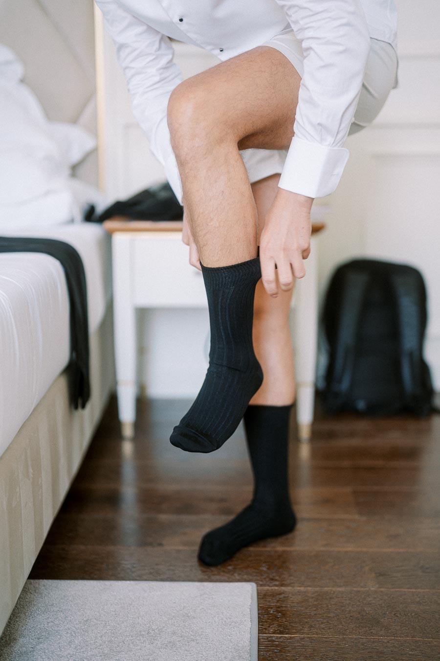 A man in formal attire sitting on a bed putting on black socks, getting ready for an event.