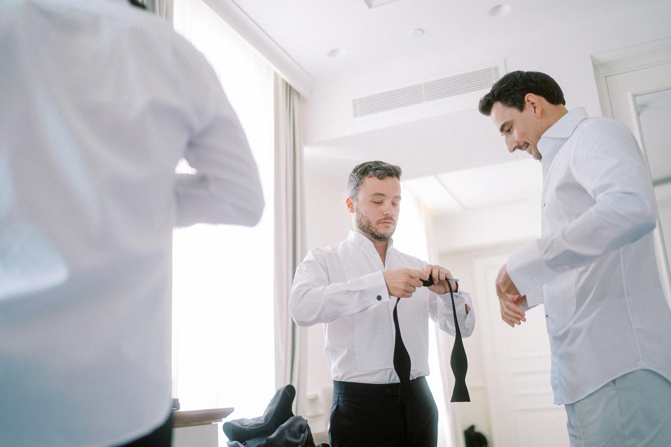 Groom and groomsmen preparing for a wedding, adjusting bow ties in a sunlit room.