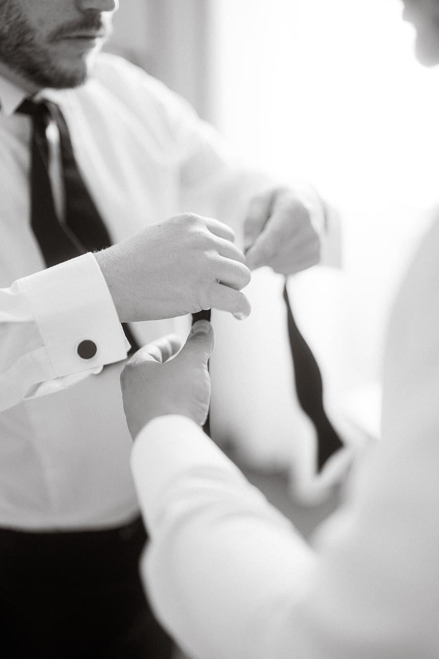 Black and white image of two men in formal attire adjusting a black tie, focusing on hands and emphasizing wedding preparation or special occasion dressing.