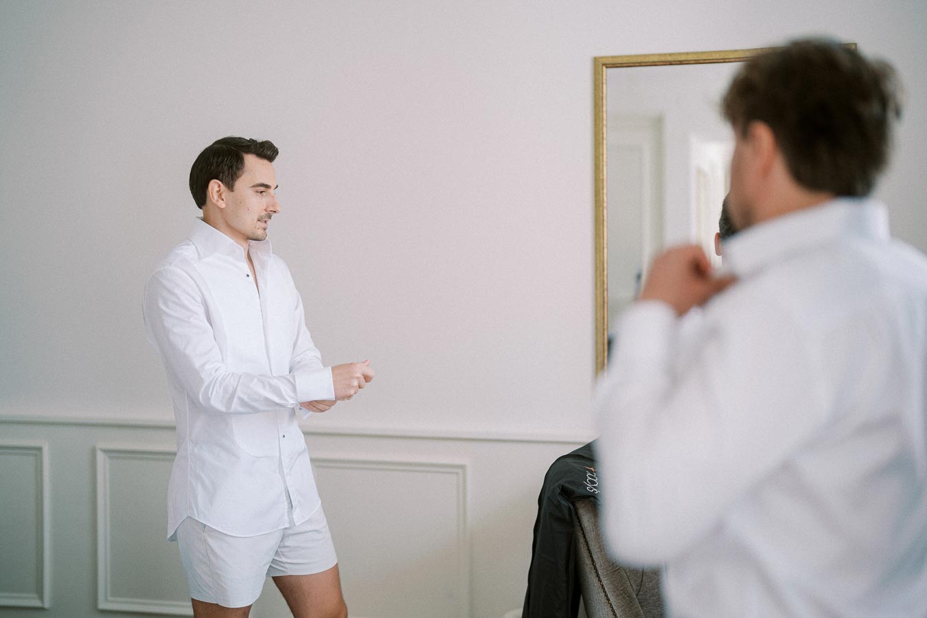 Two men getting dressed in white shirts, one adjusting his collar while standing in front of a mirror in a room with minimalistic decor.