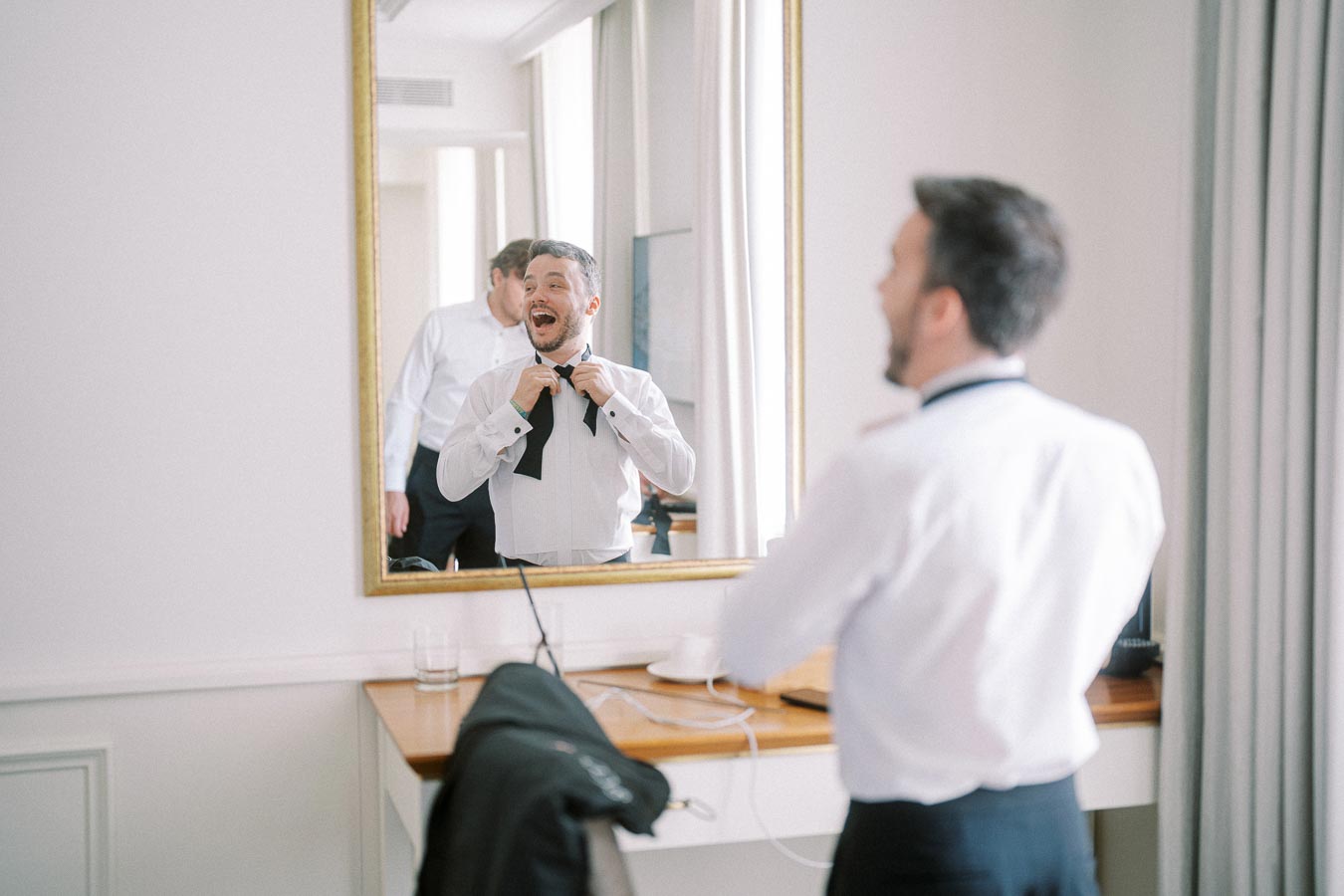 Man happily adjusting bow tie in hotel room mirror, preparing for a formal event.