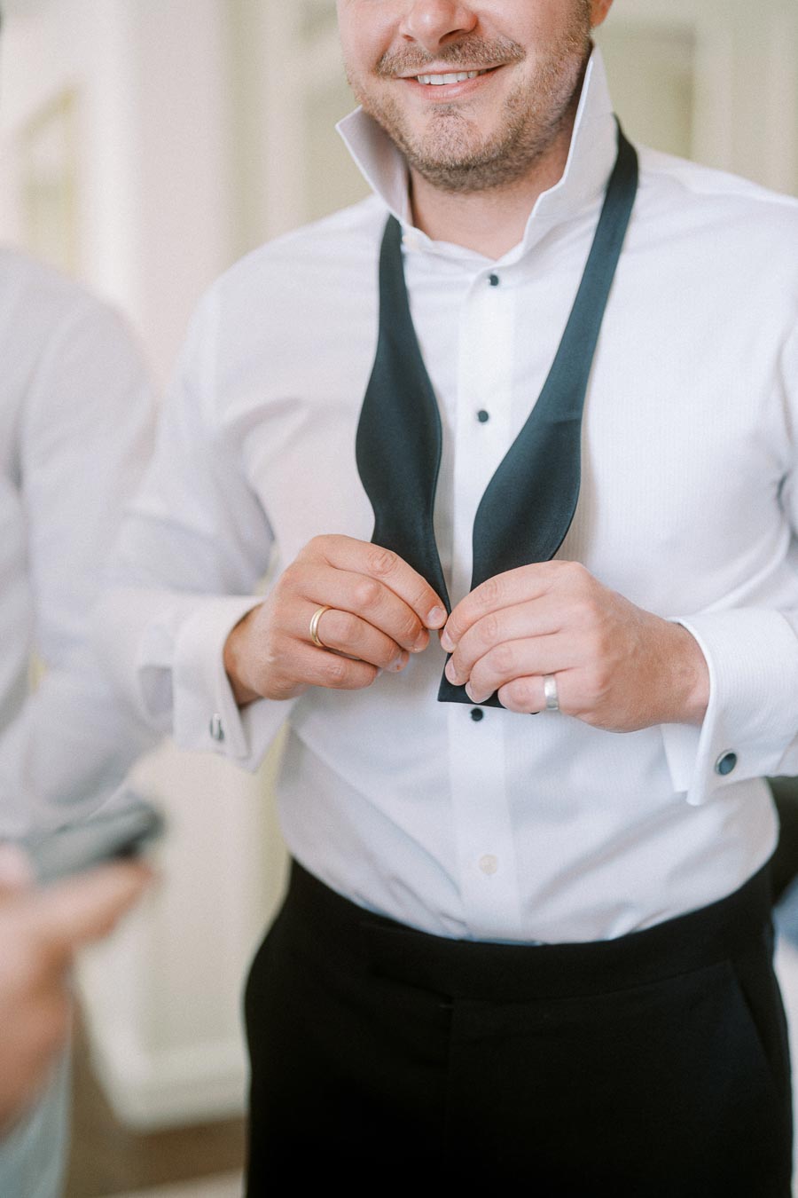 Man tying a black bow tie while wearing a formal white dress shirt, preparing for a wedding or formal event, showcasing elegant men's fashion.