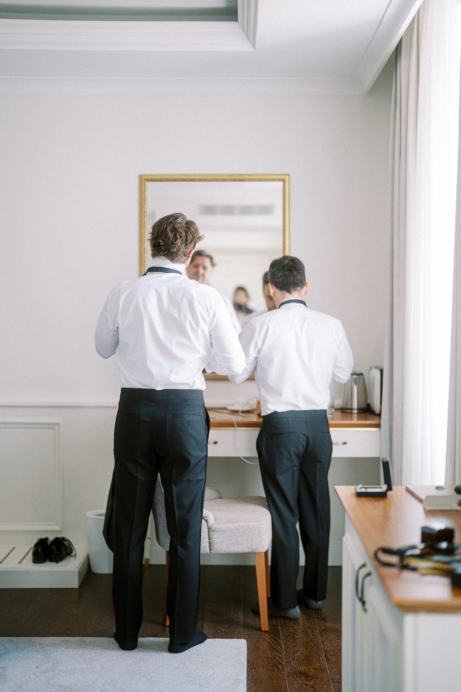Two men in white shirts and black pants adjusting their bowties in front of a mirror in a well-lit room.
