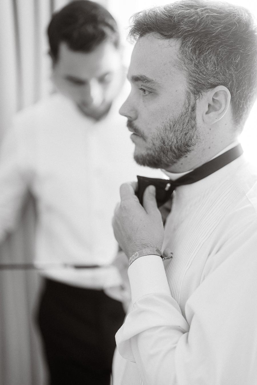 A groom adjusting his bow tie while preparing for his wedding, wearing a formal white shirt, with a groomsman assisting in the background.