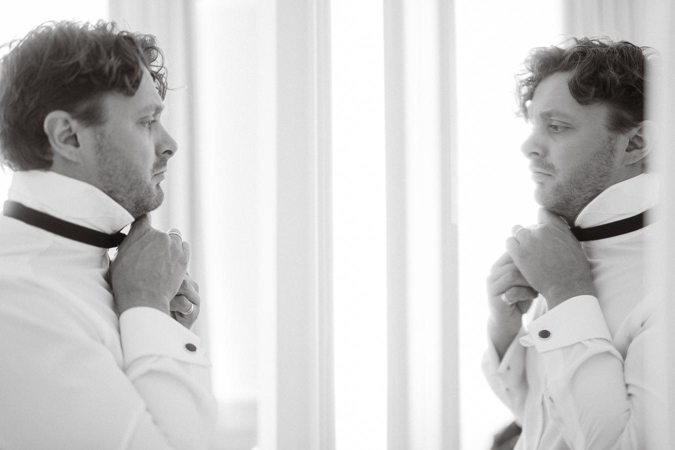 A man in formal attire adjusting his bow tie in front of a mirror, captured in a black and white setting.