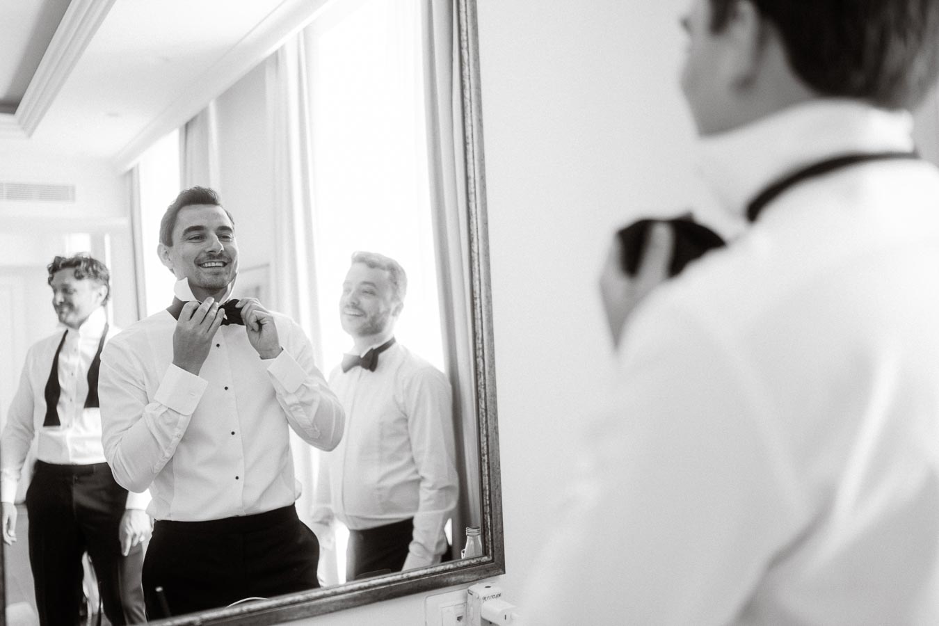 Groom and groomsmen getting ready, adjusting bow ties in a mirror, preparing for wedding ceremony.
