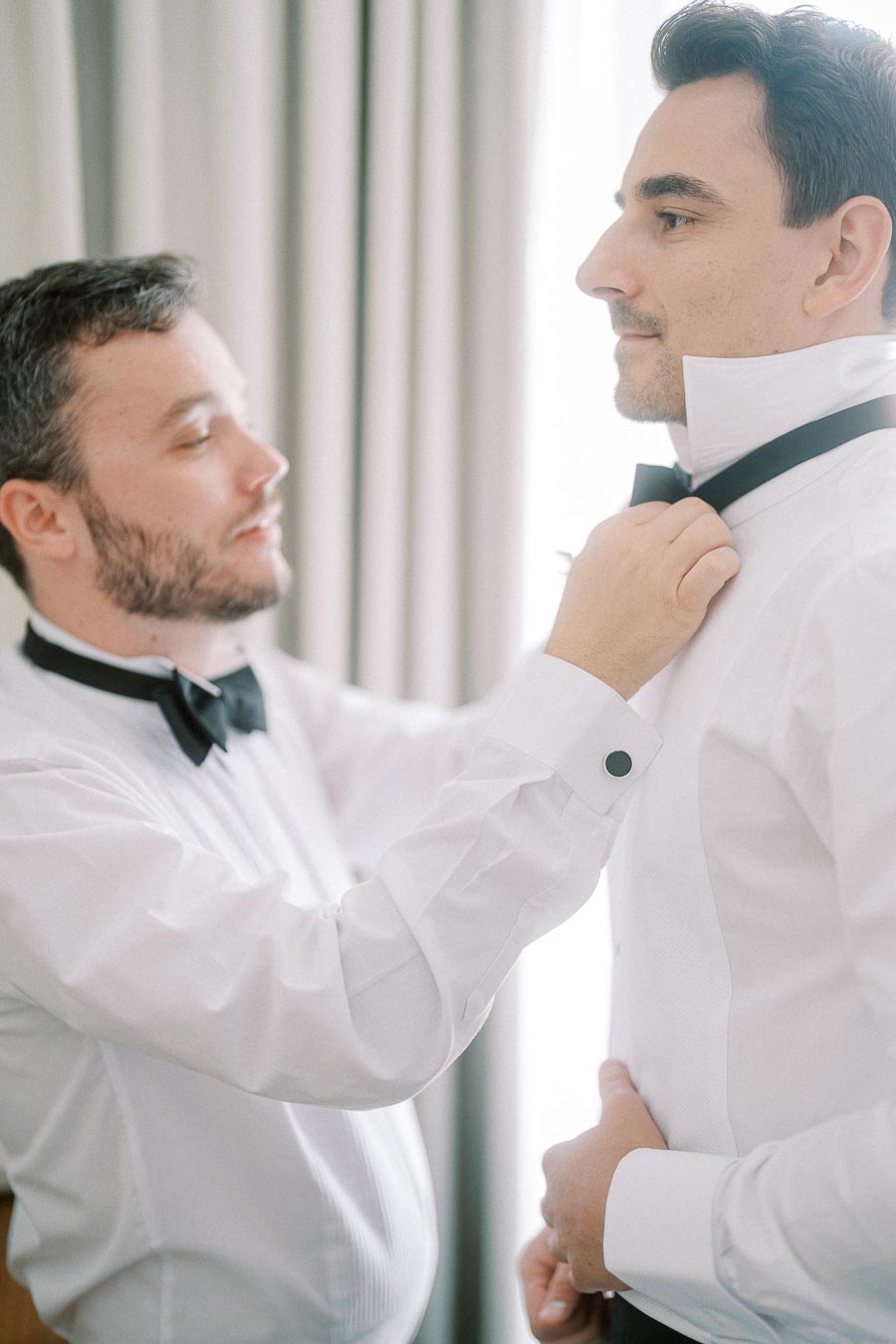 Two men in formal attire adjusting a bow tie in a bright room, one assisting the other, symbolizing preparation and camaraderie before an event.