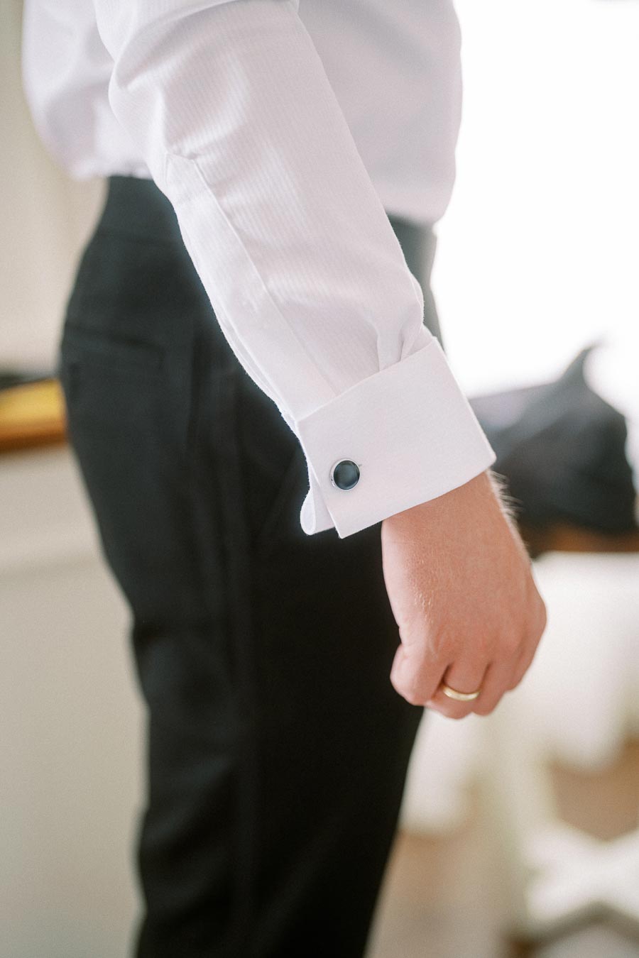 Close-up of a man's hand wearing a white dress shirt with black cufflinks and a gold wedding ring, dressed in black formal trousers, highlighting classic men's formal attire.
