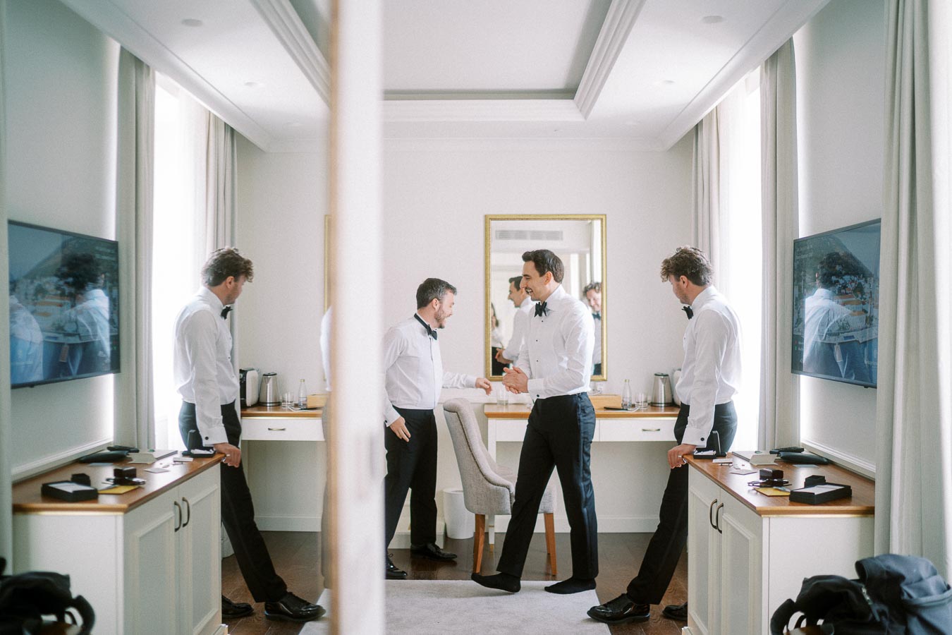 Three men in formal attire enjoy a moment in a well-lit room with elegant decor, mirrors reflecting their interactions, perfect for a luxury lifestyle or event preparation theme.