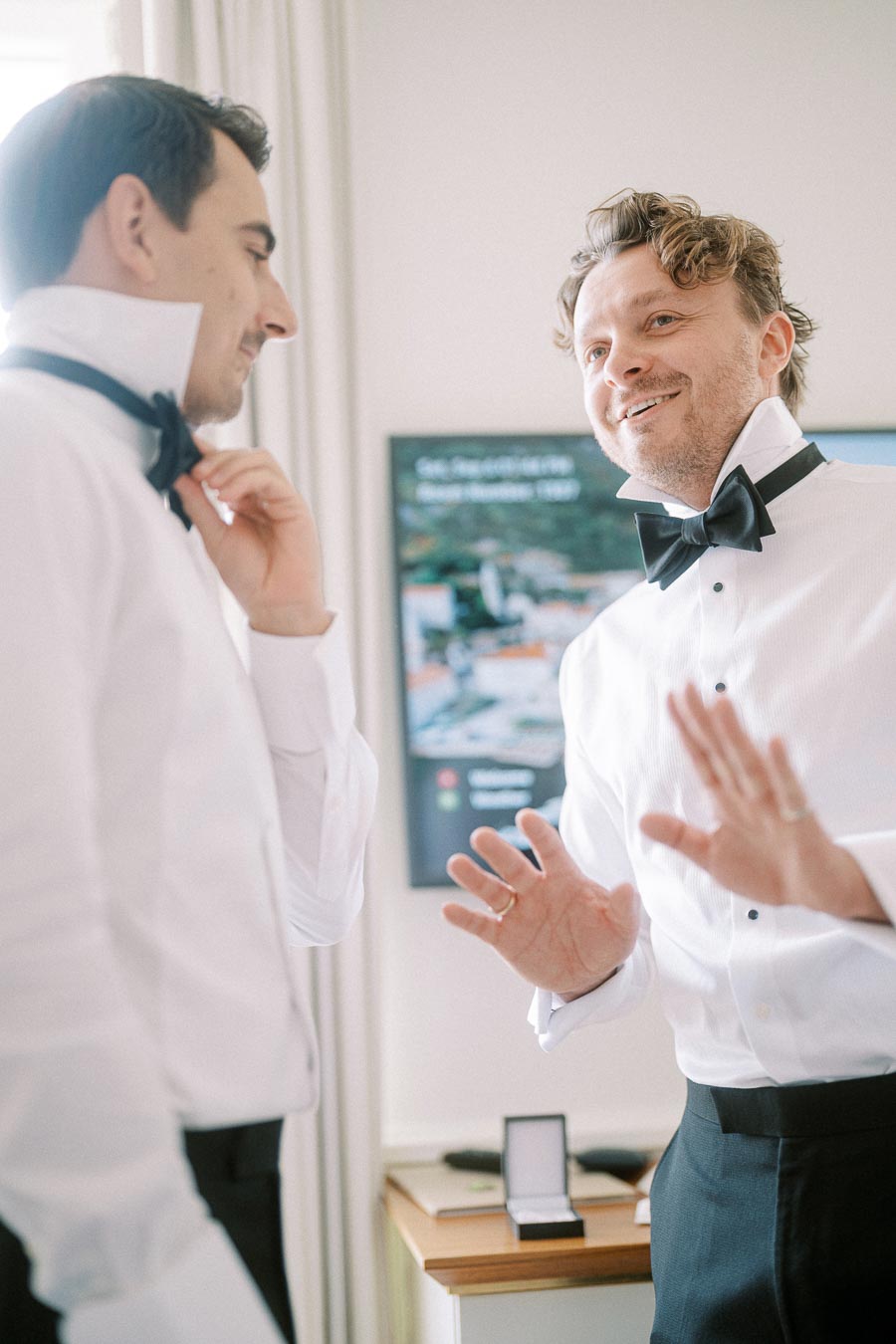 Two men in formal attire, adjusting their black bow ties and smiling in a brightly lit room, possibly preparing for a formal event or celebration.