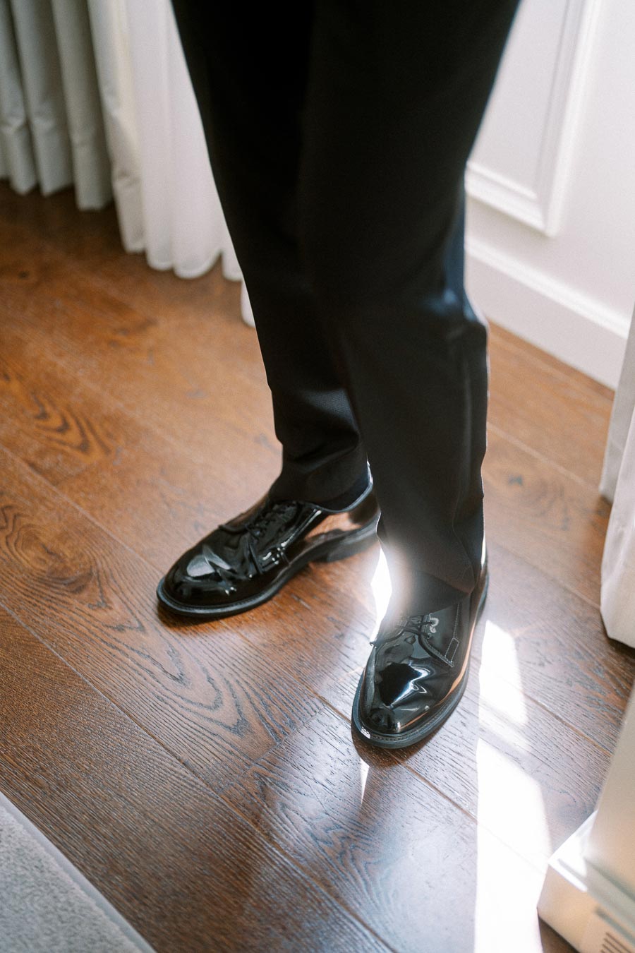 Man wearing black polished dress shoes and formal black pants standing on hardwood floor with sunlight streaming in.