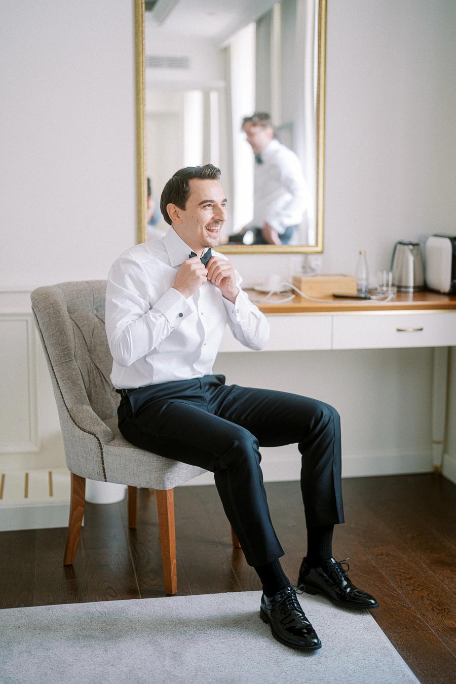 Groom preparing for wedding, adjusting bow tie while sitting on a chair in a bright, modern room with a mirror reflection.