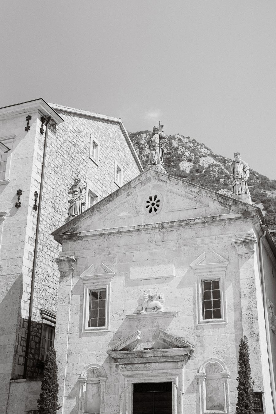 Black and white image of a historic stone church facade with sculptures, set against a mountainous backdrop in Montengro.