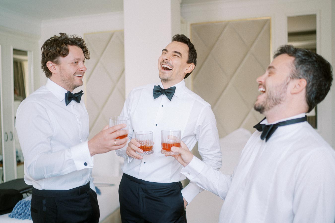 Three men in formal attire, wearing white shirts and black bow ties, laughing and toasting with drinks in a hotel room setting.