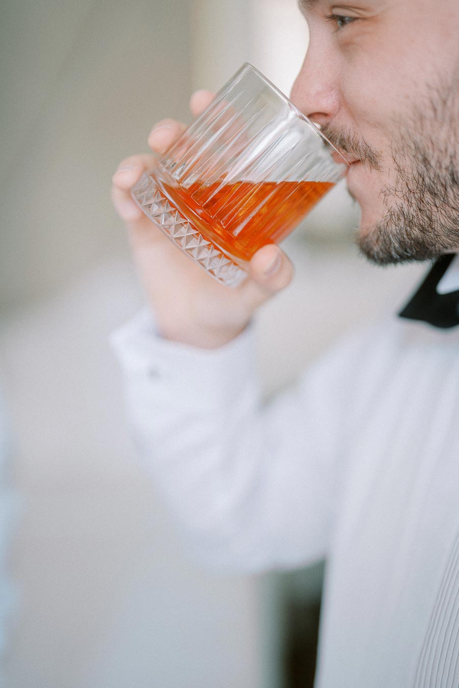Man savoring a glass of whiskey in a clear, textured glass, dressed in formal attire, highlighting relaxation and sophistication.