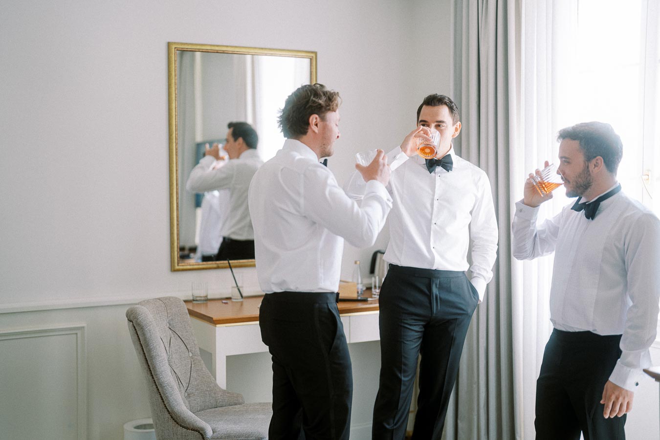 Groomsmen in formal attire enjoying drinks in a stylish room, preparing for a wedding celebration.