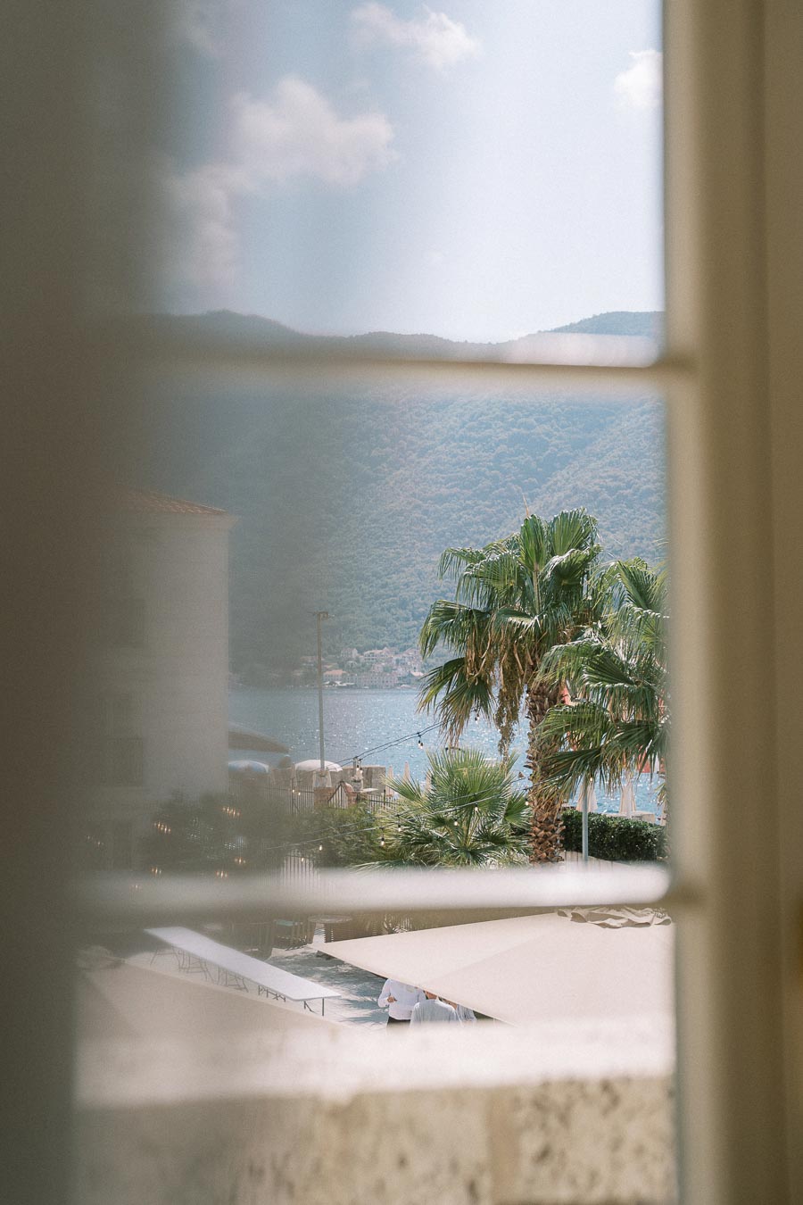 View of a picturesque coastal landscape through a window, featuring lush palm trees, a serene body of water, and distant hills under a clear sky.