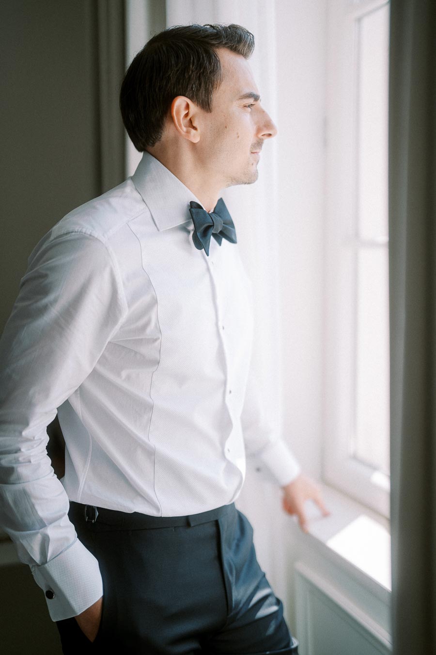 A man in a formal white shirt and black bow tie gazing out of a window, illuminated by natural light.