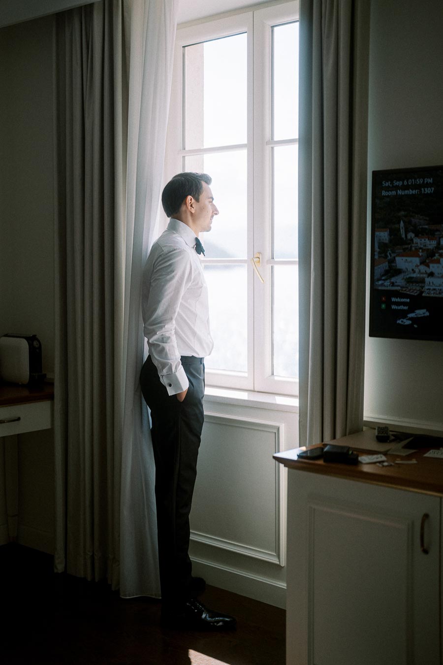 A man in a formal white shirt and black trousers stands in a hotel room, gazing out a sunlight-filled window, creating a serene and contemplative atmosphere.