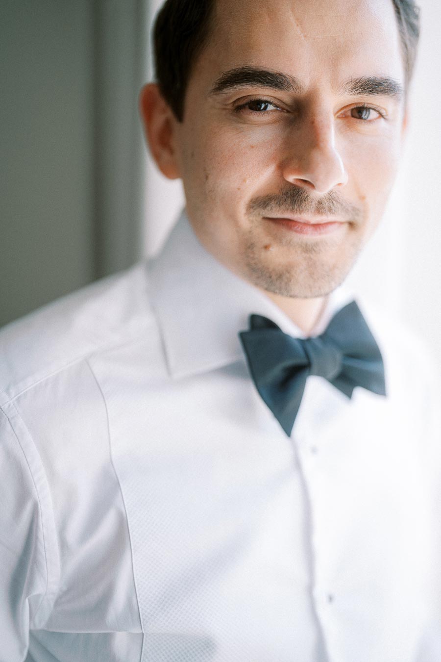 A man wearing a white dress shirt and a black bow tie, looking directly at the camera with a soft expression.