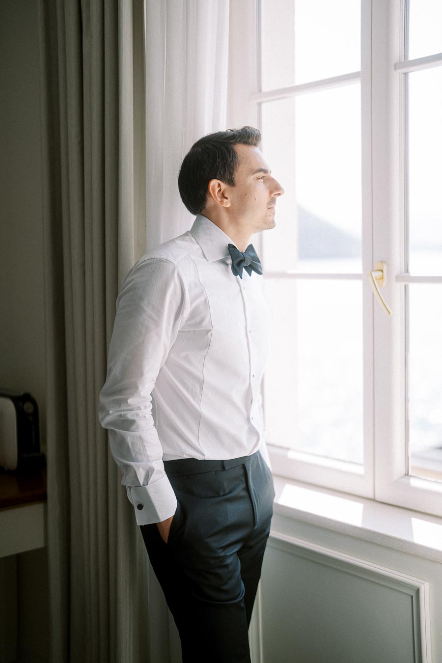 A man in a formal white shirt with a black bow tie standing by a window, looking outside, with soft light illuminating the room.