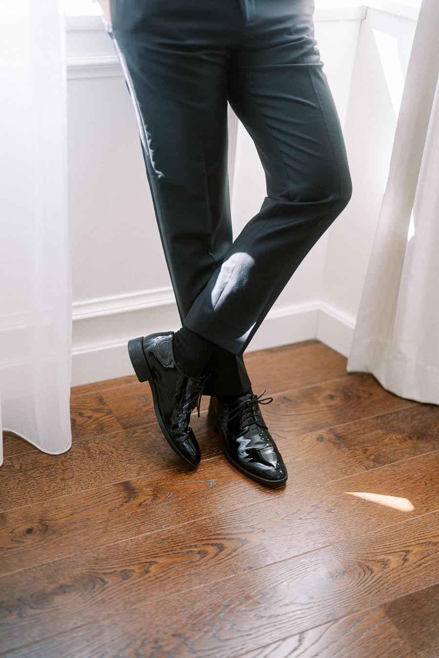 Elegant black dress shoes paired with tailored dark trousers, standing on polished hardwood flooring.