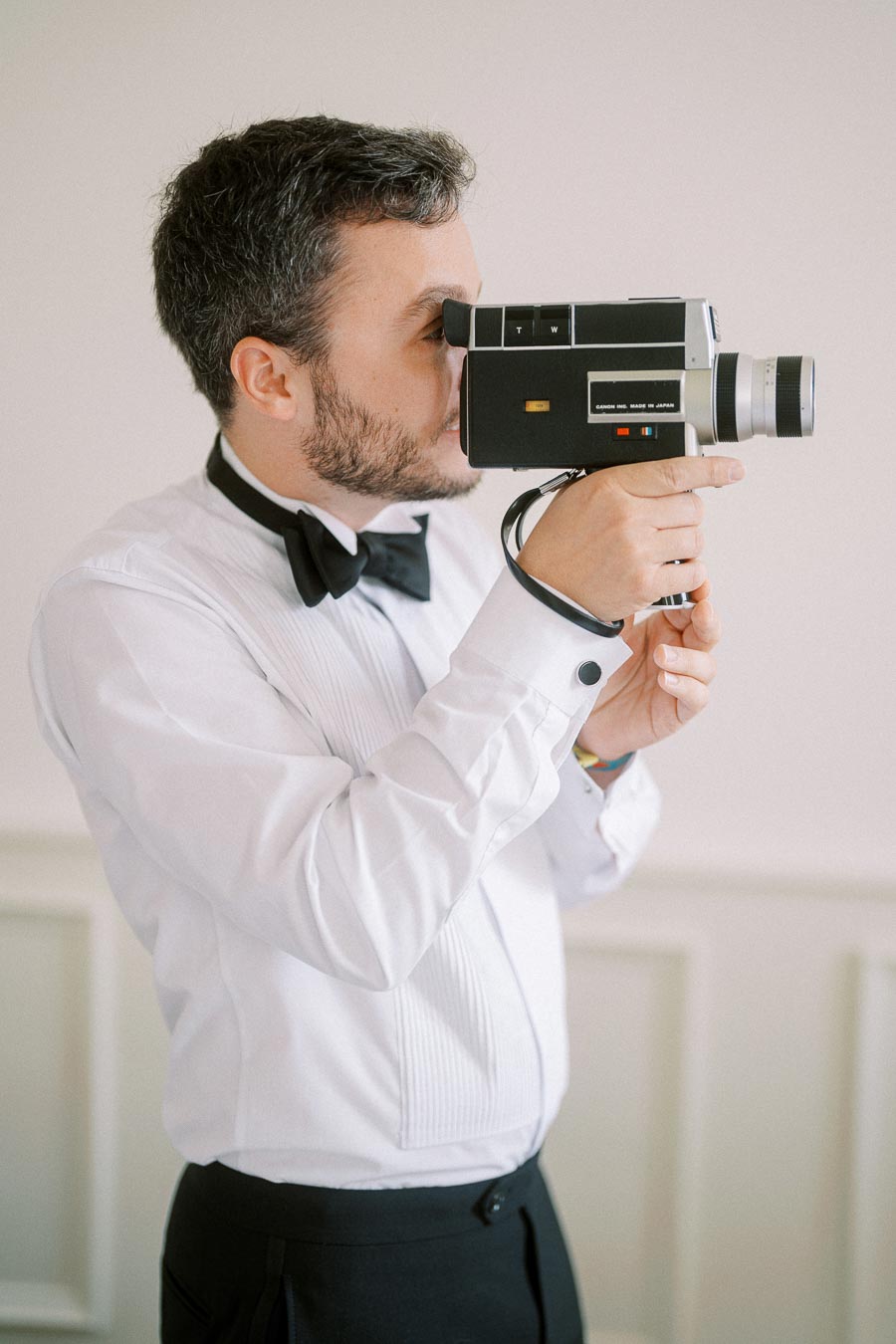 A person in formal attire, wearing a white dress shirt and black bow tie, is using a vintage film camera.