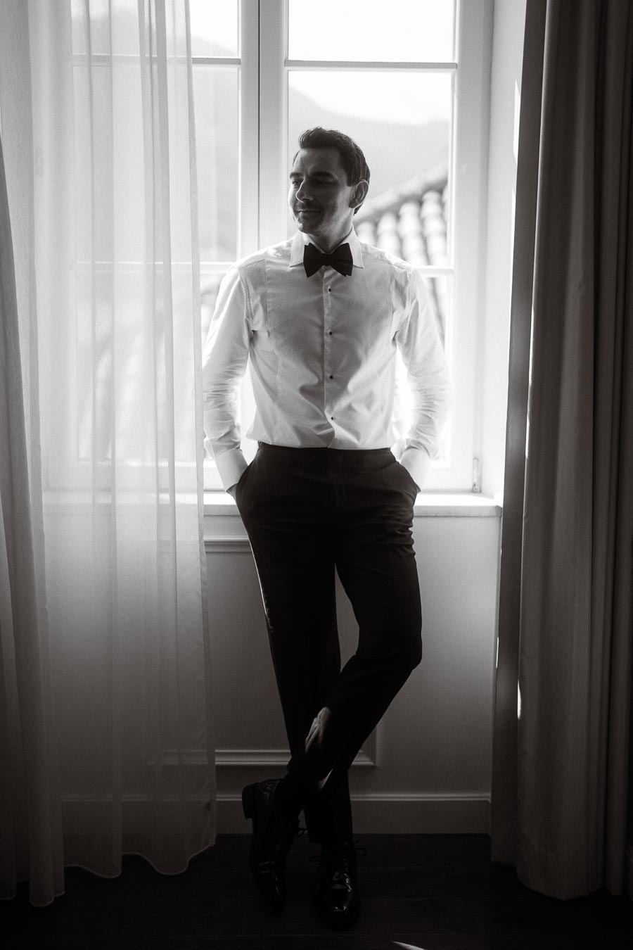 Black and white portrait of a man in a bow tie and dress shirt, standing near a window with sheer curtains.