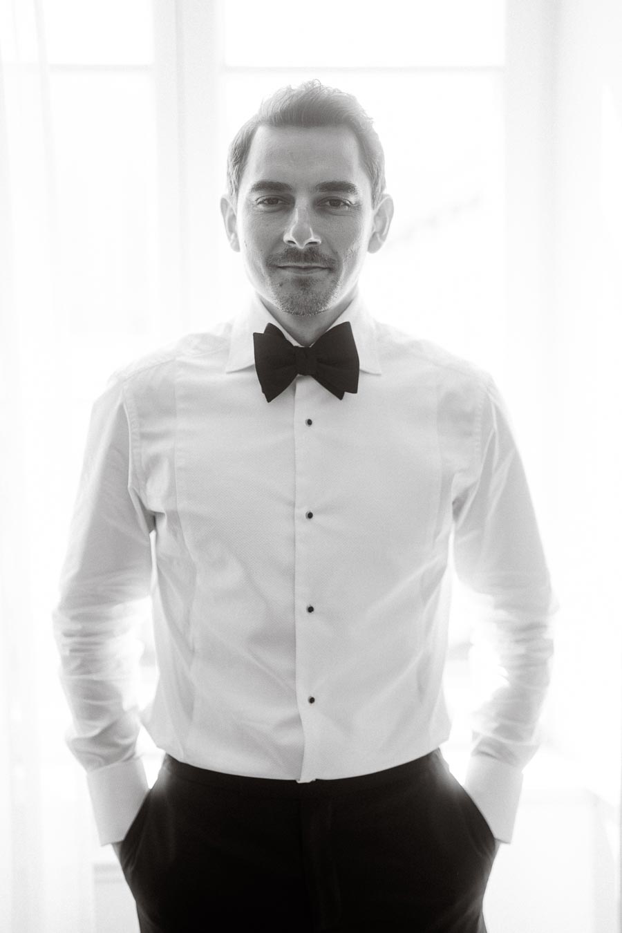 A well-dressed man in a white shirt and black bow tie stands confidently, hands in pockets, with soft light streaming in through a window behind him, creating a refined and classic look.