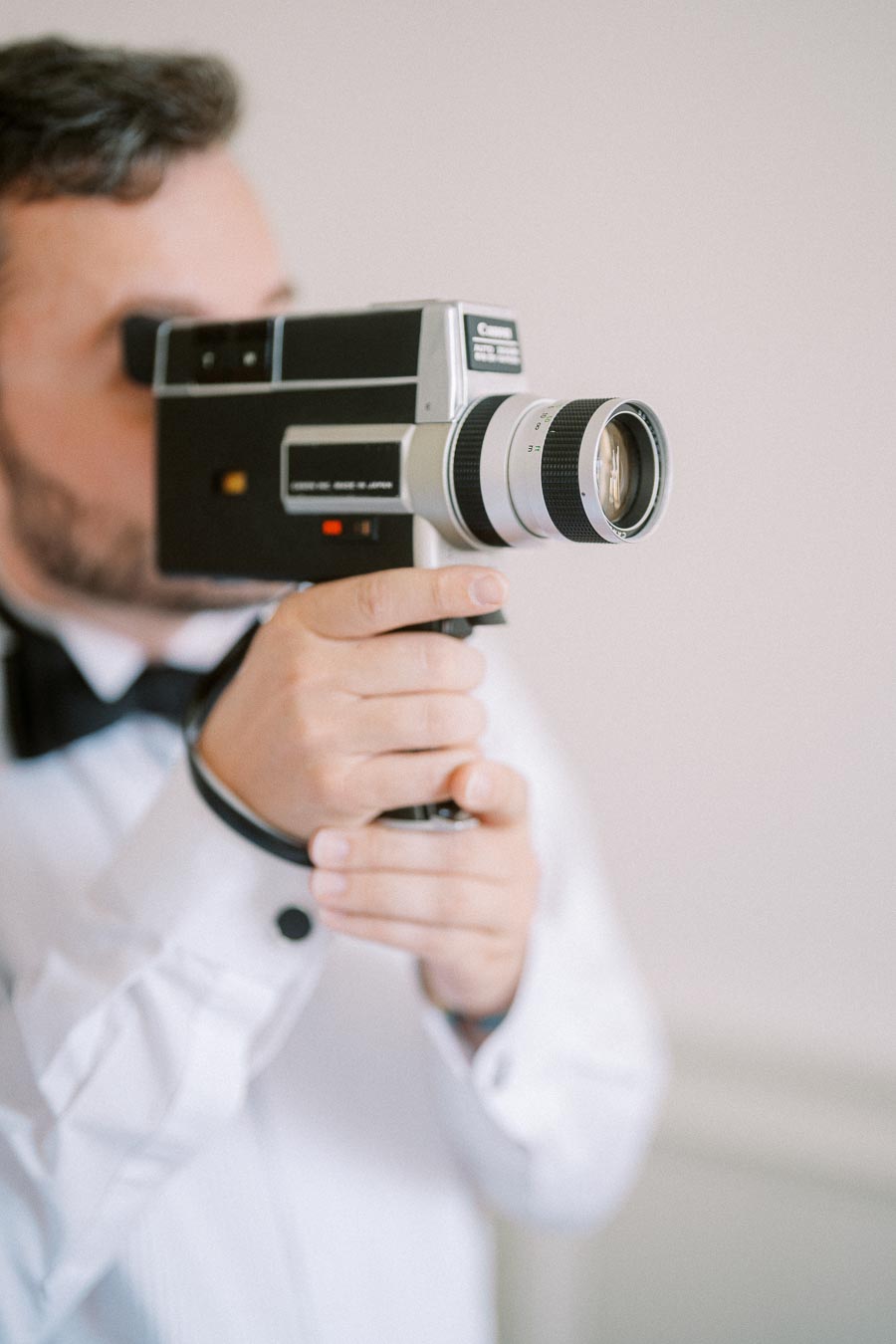 A man in a white shirt and bow tie operates a vintage video camera, focusing closely on capturing a moment.