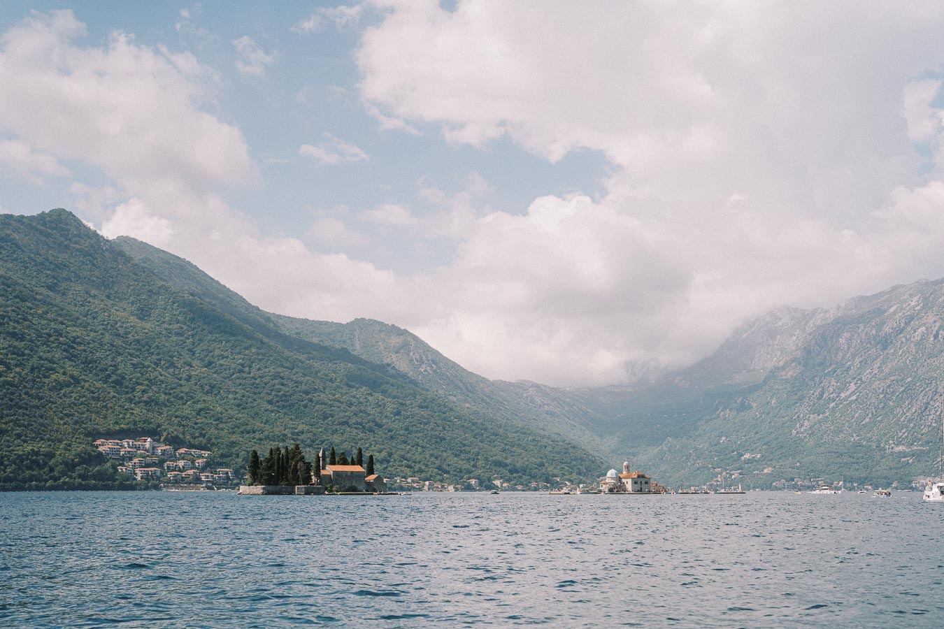 Scenic view of the Bay of Kotor in Montenegro, featuring picturesque islands with historic churches, surrounded by lush green mountains and a cloudy sky.