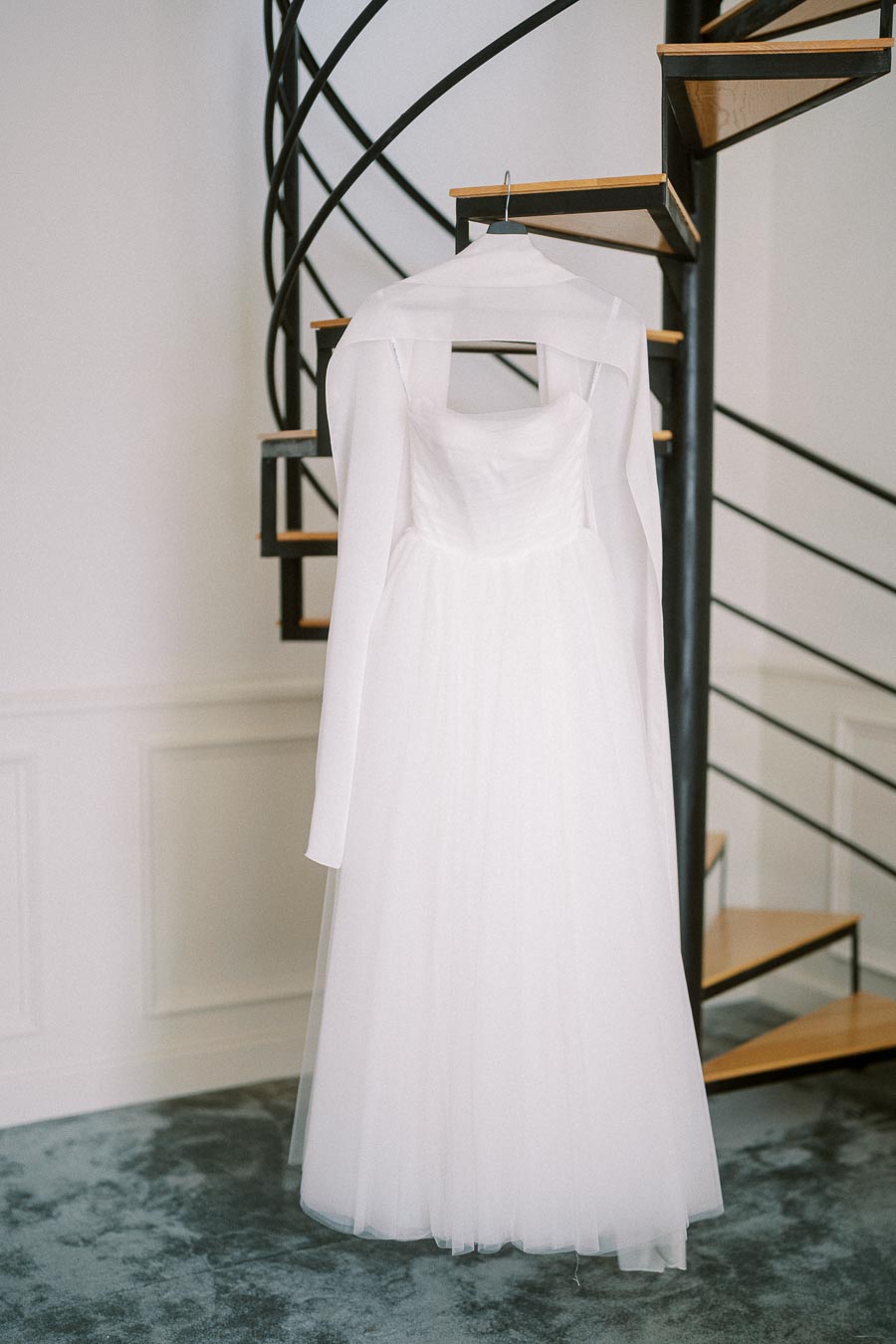 Elegant white wedding dress with long sleeves hanging in front of a stylish spiral staircase.