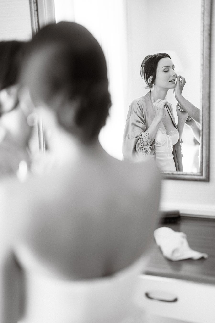 Black and white image of a bride getting ready, looking into a mirror as she applies makeup before her wedding.