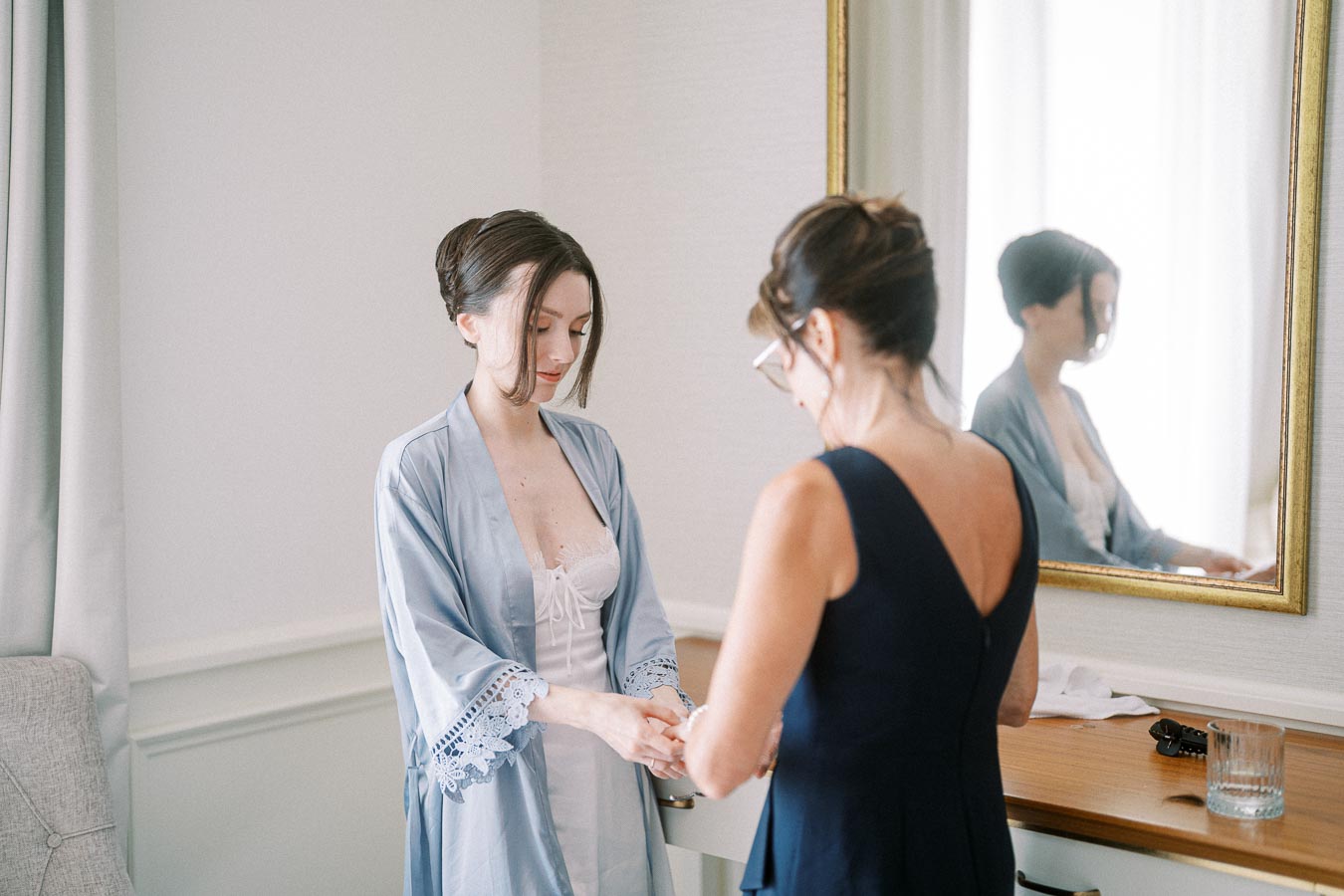 Two women preparing in a room with a large mirror. The woman on the left is wearing a blue robe and standing near the mirror, while the woman on the right is assisting her. The scene is set in a softly lit, elegant room, conveying a sense of calm and preparation.