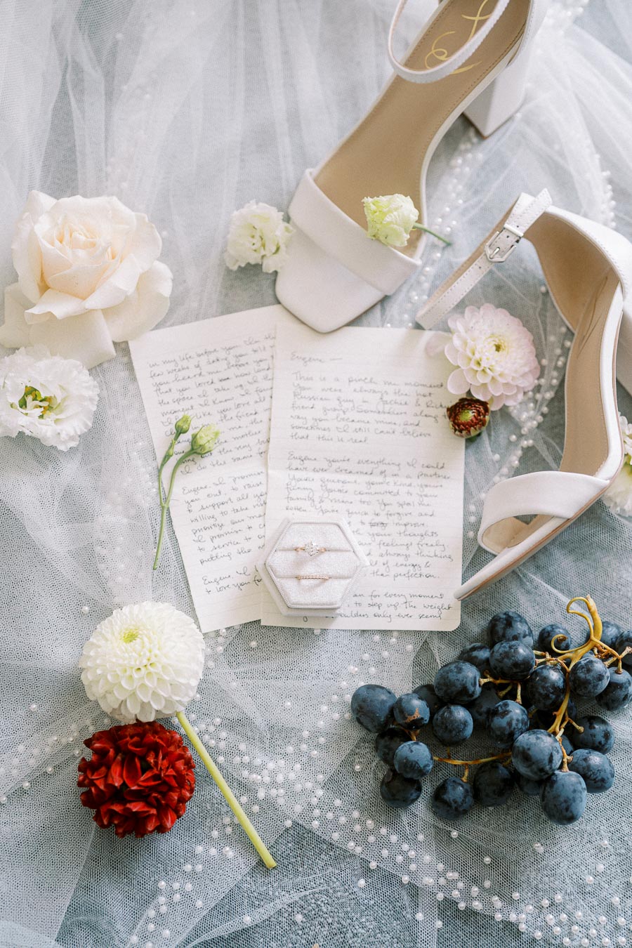 Romantic wedding flat lay featuring white high-heeled sandals, handwritten love letters, delicate flowers, an elegant ring box, and a bunch of blue grapes, all arranged on a soft, textured background. Perfect for wedding inspiration and bridal styling.