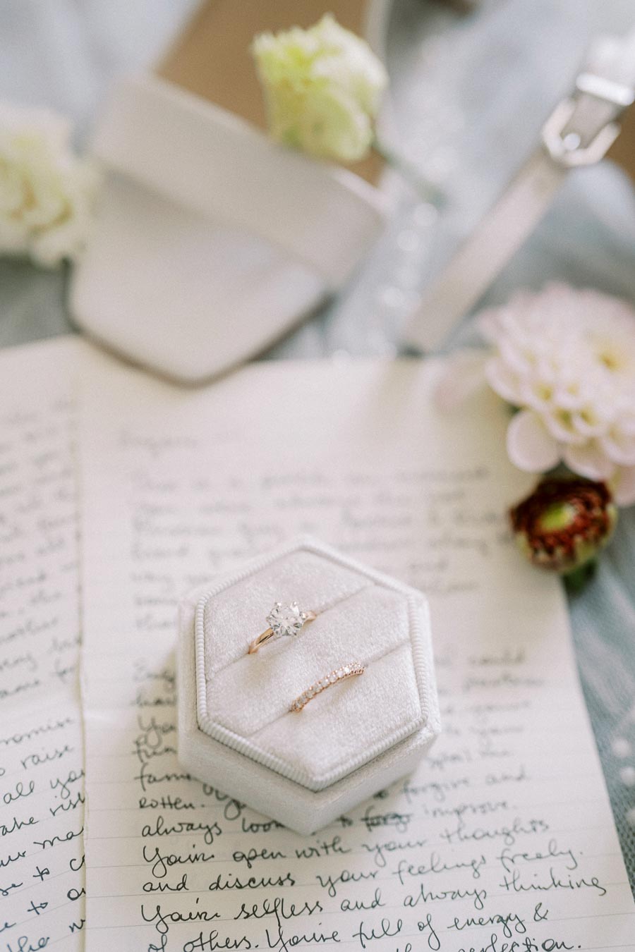 Elegant bridal jewelry featuring a diamond engagement ring and wedding band in a white velvet box, placed on handwritten letters with soft floral and shoe accents in the background, capturing a romantic and intimate wedding theme.