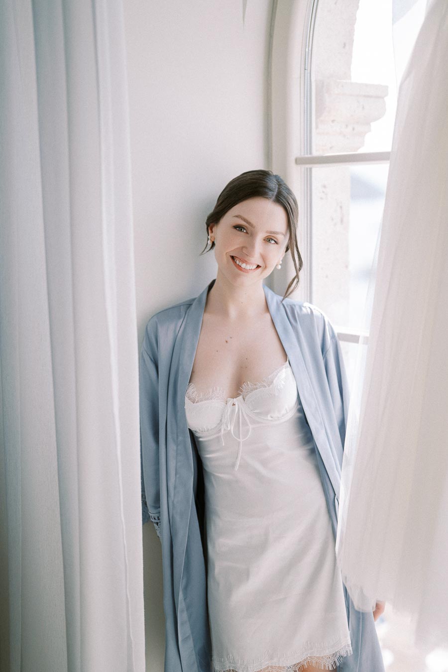 Smiling woman in white lace slip and blue robe standing by window with soft natural light