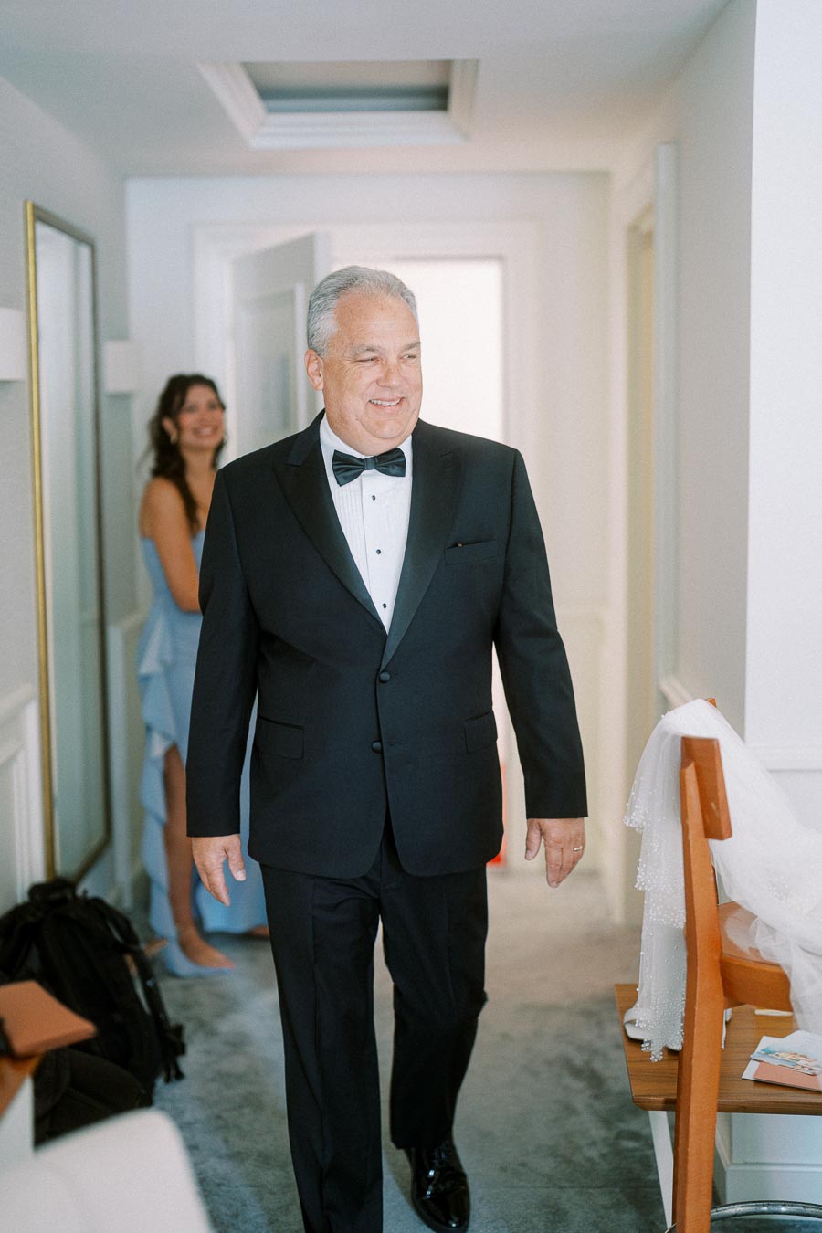 A smiling man in a black tuxedo, walking indoors, with a woman in a blue dress in the background, and a veil hanging over a chair.