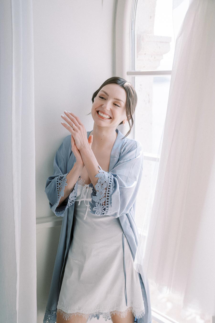 Smiling woman in a light blue lace robe standing by a window, enjoying natural light.