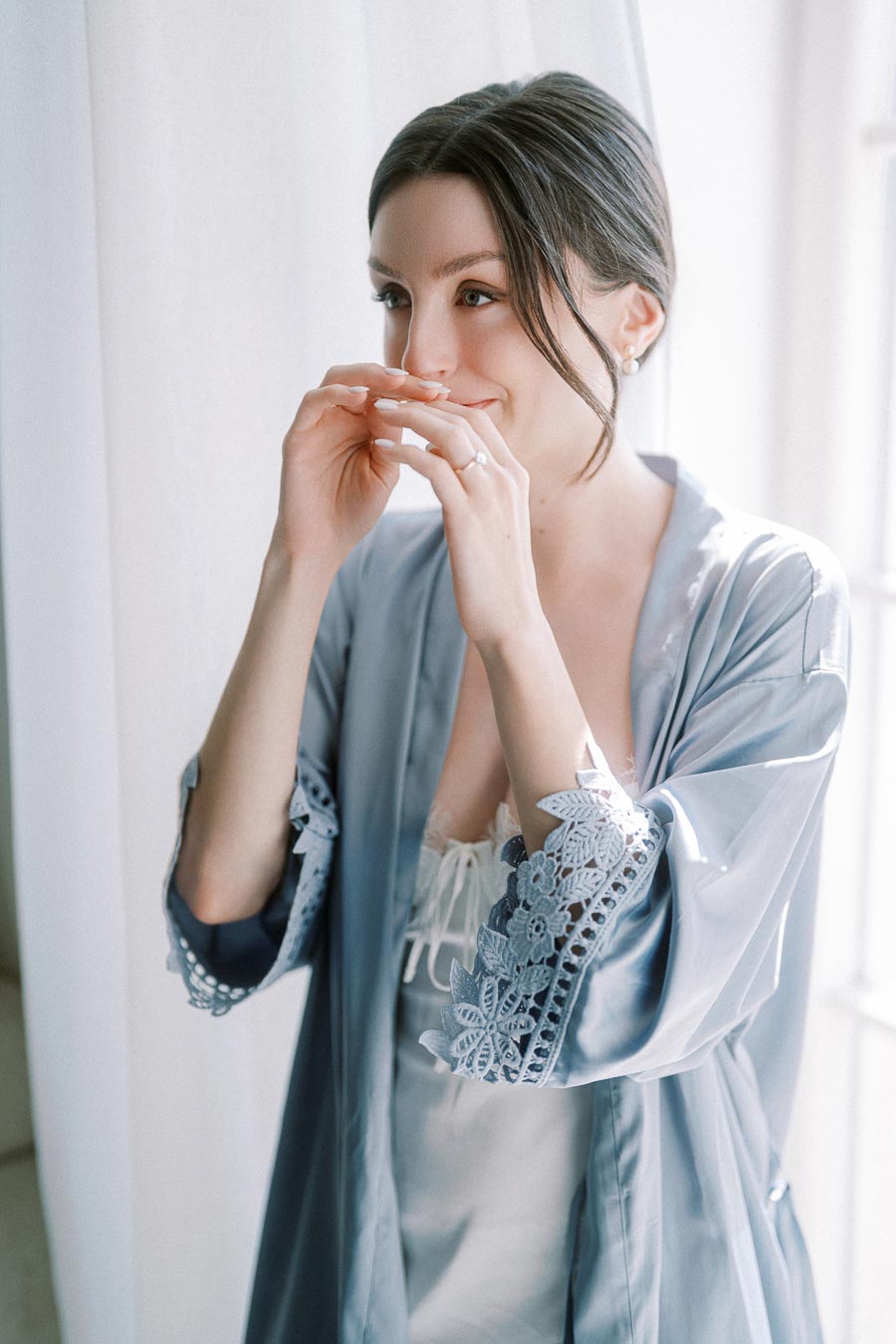A woman in a blue lace robe is standing by a window, smiling softly with her hands near her face. The natural light highlights the intricate lace details on her robe, creating an elegant and serene atmosphere.