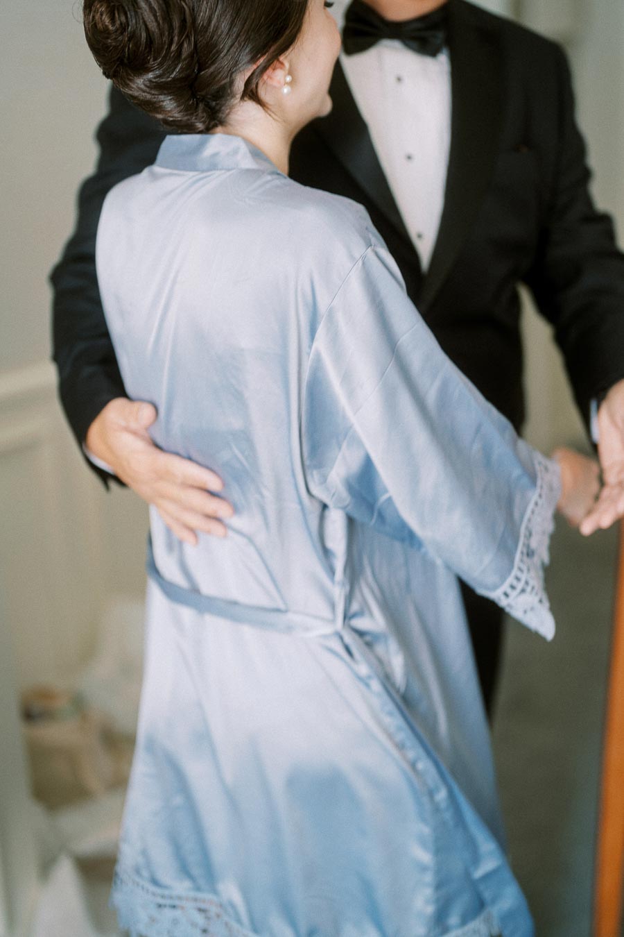 A person in a light blue robe embraces someone in a black tuxedo with a bow tie, conveying warmth and elegance.