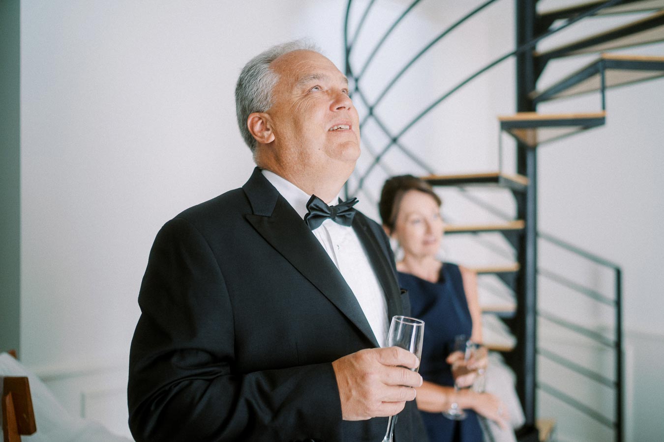 Elegant evening gathering with a man in a tuxedo holding a champagne glass, woman in formal dress in the background near a spiral staircase.