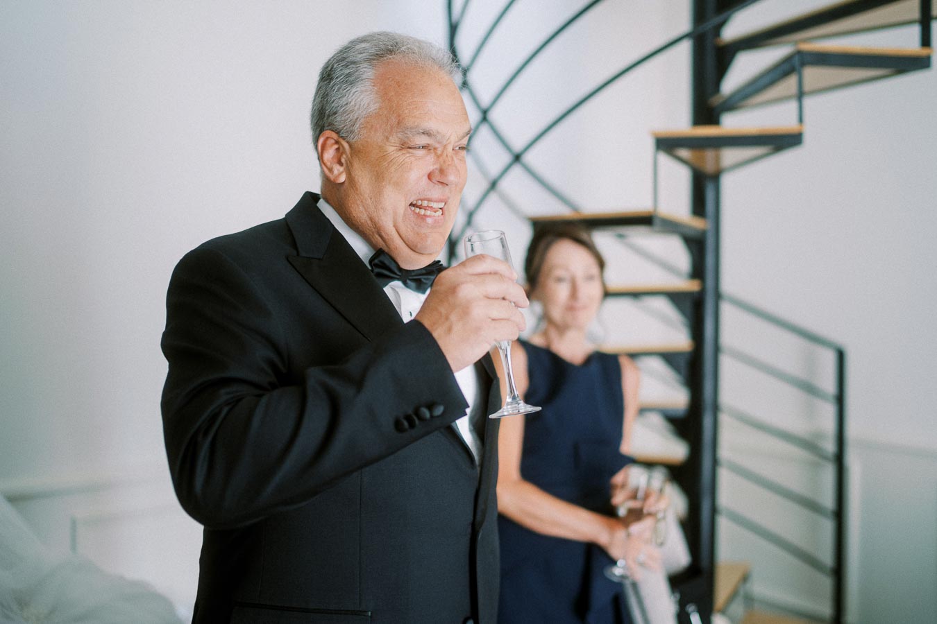 Elderly man in a tuxedo smiling and holding a champagne glass with a woman in the background, standing near a spiral staircase, celebrating a special occasion.