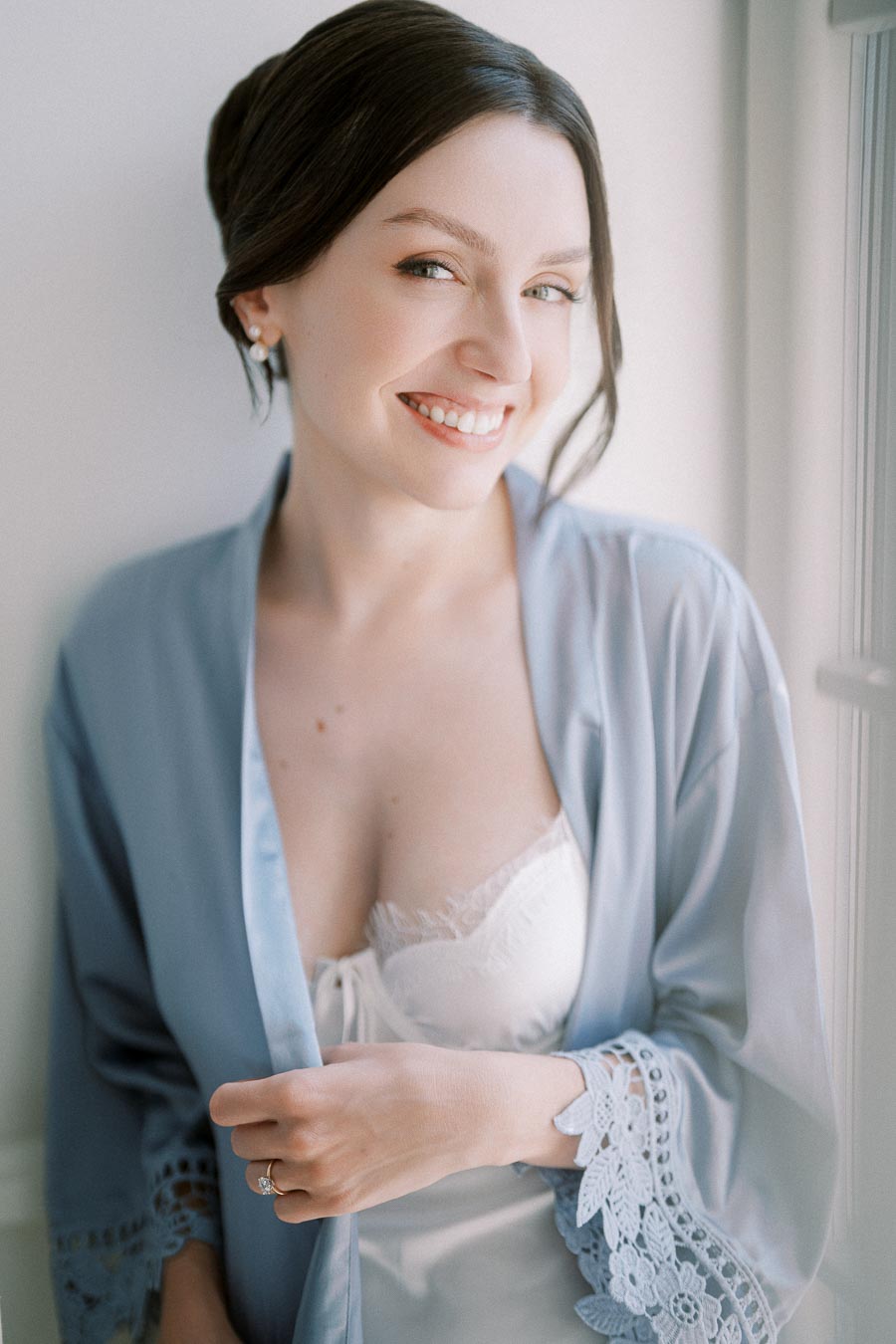 Smiling woman in a blue lace-trimmed robe, standing by a window and wearing a white lace top.