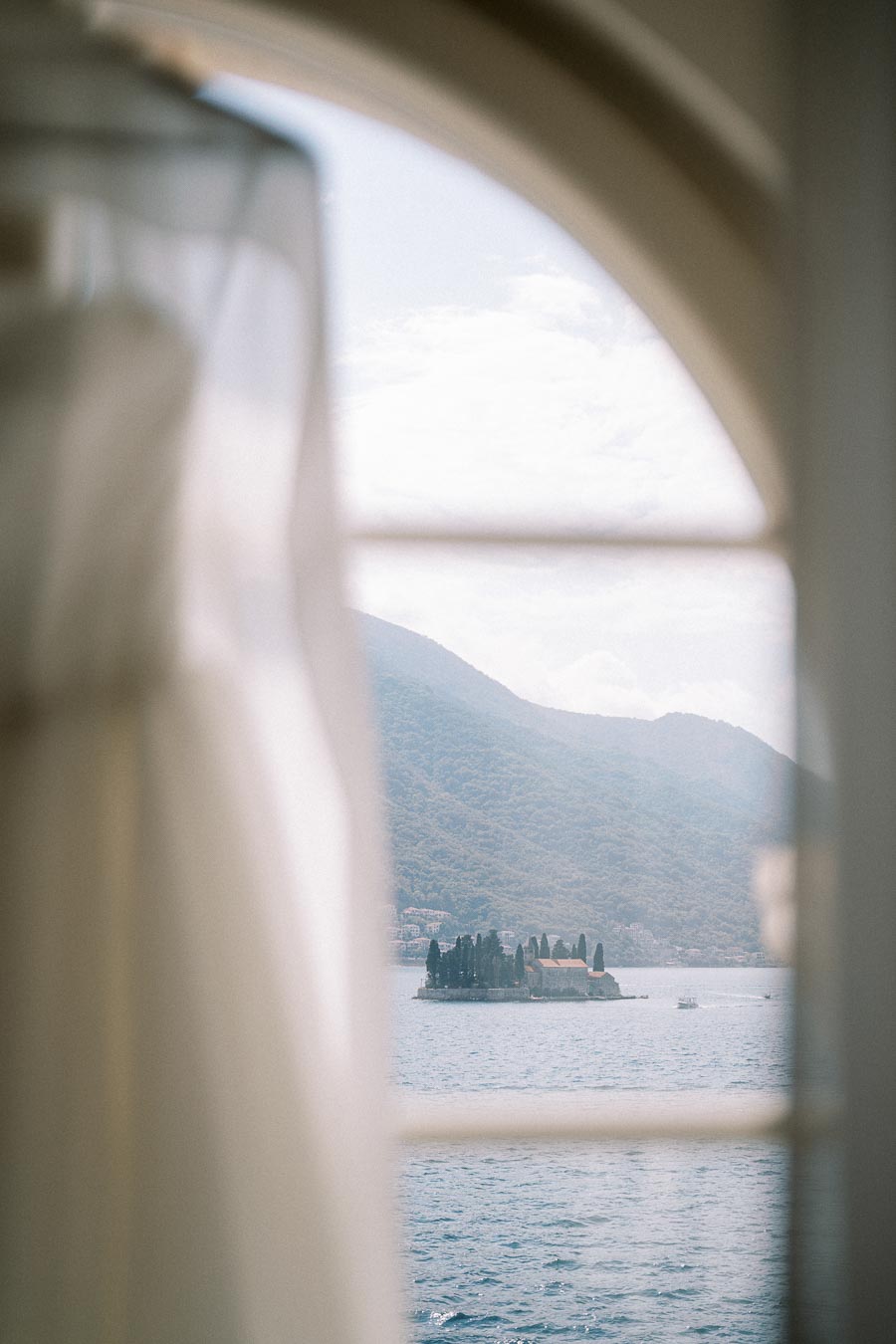 A blurred wedding dress in an elegant room frames a scenic view of an island on a tranquil lake with mountainous backdrop.