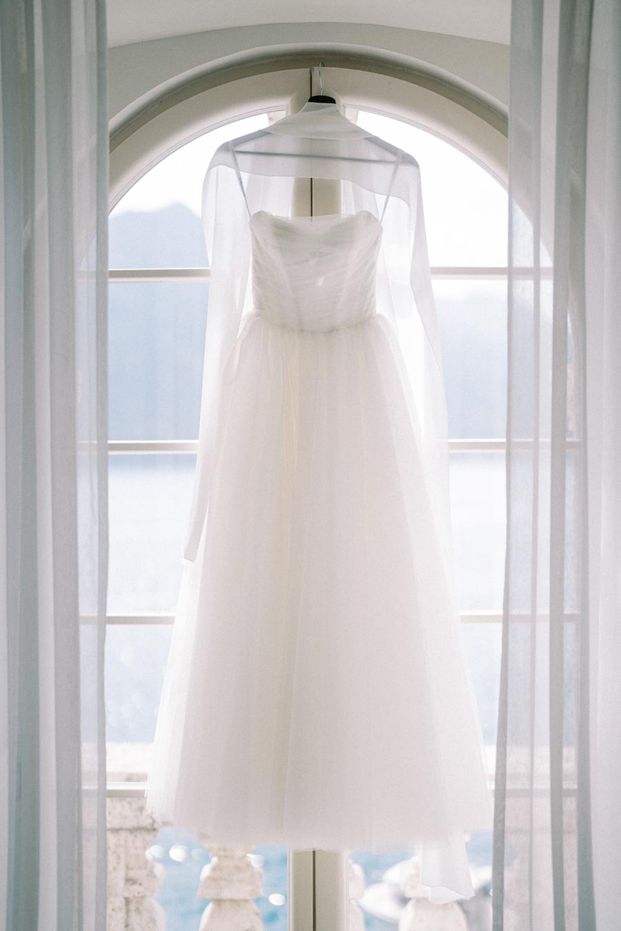 Elegant white wedding dress hanging in front of an arched window with soft, natural light.