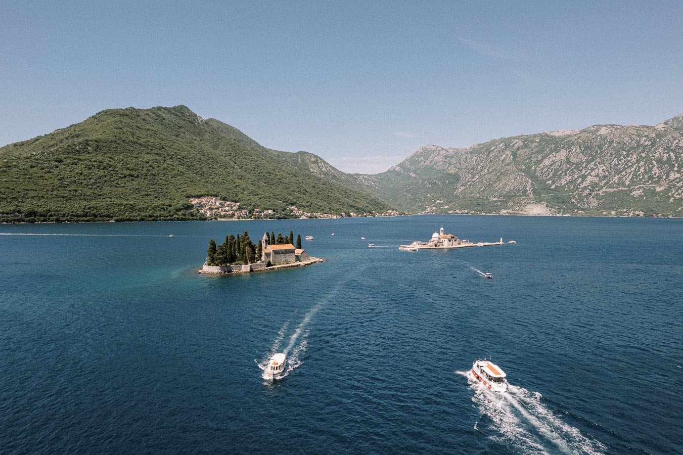 Scenic view of two small islands in the Bay of Kotor, Montenegro, featuring Saint George Island with a monastery and Our Lady of the Rocks with a church, surrounded by mountains and calm blue waters, with boats approaching the islands.