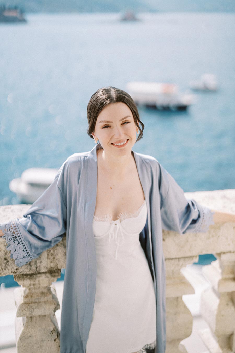 Young woman in a white dress and light blue robe smiling by a seaside balcony with boats in the background on a sunny day.
