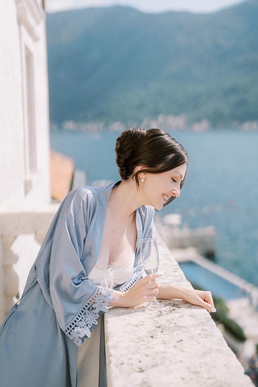 Elegant woman in a blue silk robe with lace sleeves holding an empty champagne flute, standing on a stone balcony overlooking a tranquil coastal view with mountains in the background.